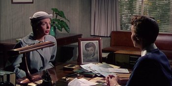 Movie still from “Magnificent Obsession” (1954), directed by Douglas Sirk – Two people sitting at a table with papers and a picture of a woman; Medium shot, Over the shoulder angle