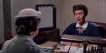 Movie still from “Magnificent Obsession” (1954), directed by Douglas Sirk – A woman sitting at a desk talking to another woman; Medium shot, Over the shoulder angle