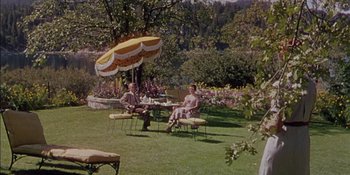 Movie still from “Magnificent Obsession” (1954), directed by Douglas Sirk – Two people sitting at a table under an umbrella in a yard; Wide shot, High angle