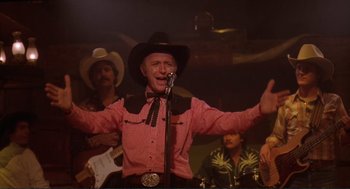 Movie still from “Making Love” (1982), directed by Arthur Hiller – A man wearing a cowboy hat singing into a microphone; Medium shot, Low angle