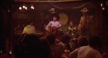 Movie still from “Making Love” (1982), directed by Arthur Hiller – A group of people sitting at a table playing guitars; Wide shot, Over the shoulder angle