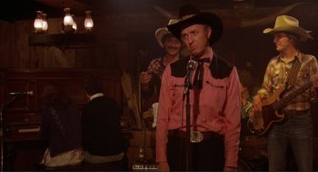 Movie still from “Making Love” (1982), directed by Arthur Hiller – A man wearing a cowboy hat and holding a microphone in front of another man; Medium shot, Over the shoulder angle