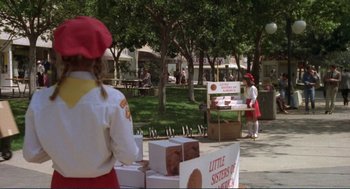 Movie still from “Making Love” (1982), directed by Arthur Hiller – A girl in a red hat is standing next to boxes of girl scout cookies; Wide shot, High angle