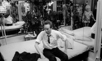Movie still from “Man Bites Dog” (1992), directed by Benoît Poelvoorde – A black and white photo of a man sitting on a bed; Medium shot, High angle