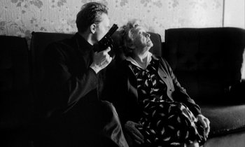 Movie still from “Man Bites Dog” (1992), directed by Benoît Poelvoorde – An older woman sitting next to a man holding a remote; Medium shot, Over the shoulder angle