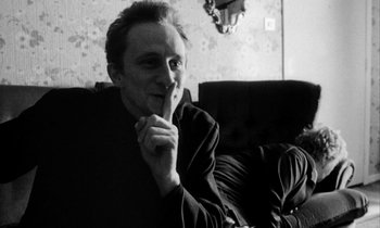 Movie still from “Man Bites Dog” (1992), directed by Benoît Poelvoorde – A man sitting on a couch with his finger on his lips; Close Up shot, Over the shoulder angle