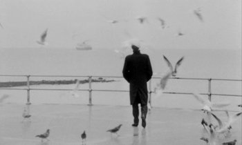 Movie still from “Man Bites Dog” (1992), directed by Benoît Poelvoorde – A man standing in the rain next to the ocean; Wide shot, Low angle
