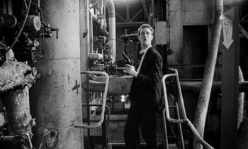 Movie still from “Man Bites Dog” (1992), directed by Benoît Poelvoorde – A man in a suit and tie standing next to pipes; Medium shot, Low angle