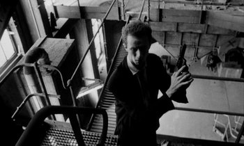 Movie still from “Man Bites Dog” (1992), directed by Benoît Poelvoorde – A black and white photo of a man holding a gun; Medium shot, Low angle