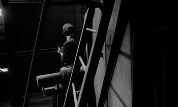Movie still from “Man Bites Dog” (1992), directed by Benoît Poelvoorde – A man sitting on a chair in a dark room; Wide shot, High angle