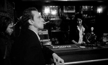 Movie still from “Man Bites Dog” (1992), directed by Benoît Poelvoorde – A man sitting at a bar smoking a cigarette and a woman sitting at a bar; Medium shot, Low angle