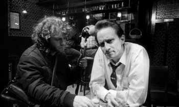 Movie still from “Man Bites Dog” (1992), directed by Benoît Poelvoorde – A man sitting at a table with a microphone in front of another man; Close Up shot, Over the shoulder angle