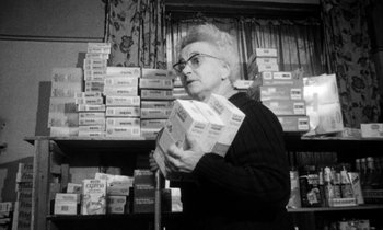 Movie still from “Man Bites Dog” (1992), directed by Benoît Poelvoorde – An older woman holding boxes in a room full of boxes; Medium shot, Low angle