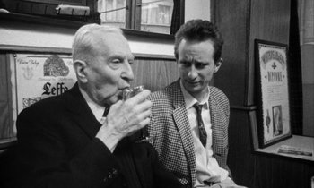 Movie still from “Man Bites Dog” (1992), directed by Benoît Poelvoorde – Two men sitting next to each other and one of them is holding a drink; Medium shot, Over the shoulder angle