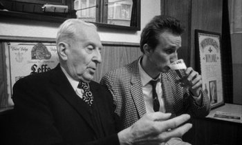 Movie still from “Man Bites Dog” (1992), directed by Benoît Poelvoorde – Two men sitting at a table with a drink in their hands; Medium shot, Low angle