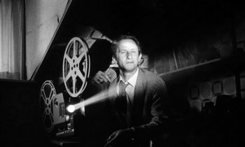 Movie still from “Man Bites Dog” (1992), directed by Benoît Poelvoorde – A black and white photo of a man in a suit and tie in front of an old movie projector; Medium shot, Low angle
