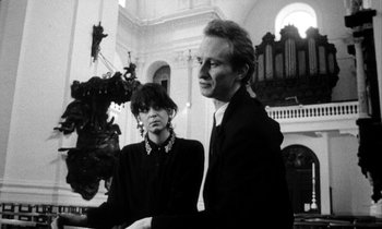 Movie still from “Man Bites Dog” (1992), directed by Benoît Poelvoorde – A man and a woman standing next to each other in front of an organ; Medium shot, Low angle