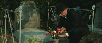 Movie still from “Manon of the Spring” (1986), directed by Claude Berri – A man in a black suit and black hat kneeling over a flower arrangement in front of a grave; Close Up shot, High angle
