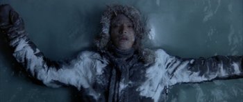 Movie still from “Map of the Human Heart” (1992), directed by Vincent Ward – A man is laying in the snow wearing a coat and hat; Medium shot, Overhead angle