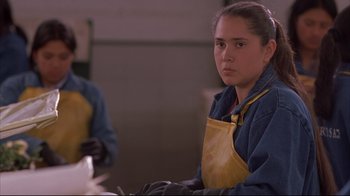 Movie still from “Maria Full of Grace” (2004), directed by Joshua Marston – A woman wearing an apron and gloves is sitting in a room; Medium shot, Low angle
