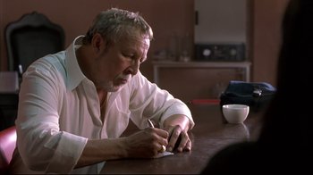 Movie still from “Maria Full of Grace” (2004), directed by Joshua Marston – An older man writing on a piece of paper on top of a wooden table; Close Up shot, Over the shoulder angle