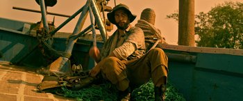 Movie still from “Extraction” (2020), directed by Sam Hargrave – A man sitting on top of a pile of rope; Medium shot, Low angle