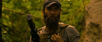 Movie still from “Extraction” (2020), directed by Sam Hargrave – A man with a beard holding a rifle in a forest; Close Up shot, Over the shoulder angle