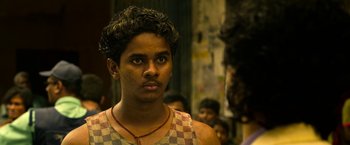 Movie still from “Extraction” (2020), directed by Sam Hargrave – A young man in a checkered tank top looks into the camera; Close Up shot, Over the shoulder angle