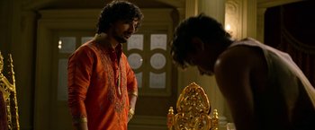 Movie still from “Extraction” (2020), directed by Sam Hargrave – A man in an orange shirt standing in front of a chair; Medium shot, Over the shoulder angle