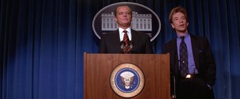 Movie still from “Mars Attacks!” (1996), directed by Tim Burton – A man standing at a podium in front of a sign; Medium shot, Low angle