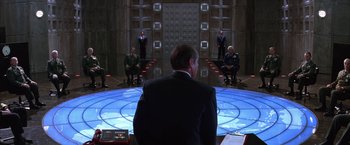 Movie still from “Mars Attacks!” (1996), directed by Tim Burton – A group of people sitting in a room with a man in a suit; Wide shot, High angle
