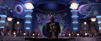 Movie still from “Mars Attacks!” (1996), directed by Tim Burton – A man standing at a table in front of a group of people; Wide shot, Over the shoulder angle