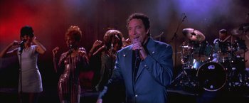 Movie still from “Mars Attacks!” (1996), directed by Tim Burton – A man singing into a microphone in front of a group of people; Medium shot, Low angle