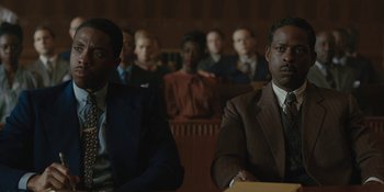 Movie still from “Marshall” (2017), directed by Reginald Hudlin – Two men in suits sitting in front of a crowd of onlookers; Medium shot, High angle
