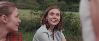Movie still from “Martha Marcy May Marlene” (2011), directed by Sean Durkin – A young woman sitting in the grass smiling for the camera; Close Up shot, Over the shoulder angle