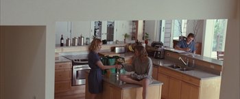 Movie still from “Martha Marcy May Marlene” (2011), directed by Sean Durkin – Two people sitting at a kitchen counter and one is holding a glass of wine; Wide shot, High angle