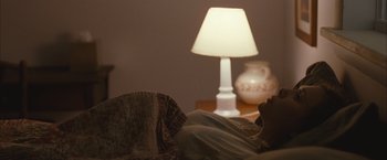 Movie still from “Martha Marcy May Marlene” (2011), directed by Sean Durkin – A person laying in a bed next to a table with a lamp; Medium shot, Over the shoulder angle