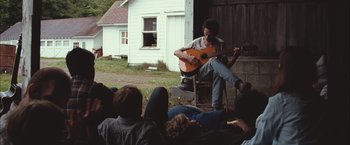 Movie still from “Martha Marcy May Marlene” (2011), directed by Sean Durkin – A man sitting on a chair playing a guitar; Wide shot, Over the shoulder angle