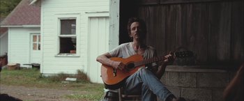 Movie still from “Martha Marcy May Marlene” (2011), directed by Sean Durkin – A man sitting in a chair playing a guitar; Medium shot, Low angle