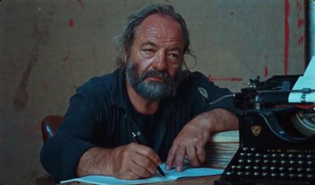 Movie still from “Martin Eden” (2019), directed by Pietro Marcello – An older man writing on a piece of paper at a desk; Medium shot, Low angle