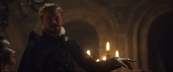 Movie still from “Mary Queen of Scots” (2018), directed by Josie Rourke – A man in a black and white outfit standing in front of candles; Medium shot, Over the shoulder angle
