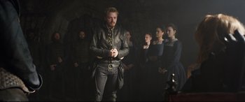 Movie still from “Mary Queen of Scots” (2018), directed by Josie Rourke – A group of people standing in a dark room; Medium shot, Over the shoulder angle