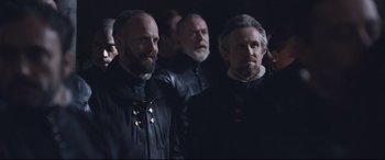 Movie still from “Mary Queen of Scots” (2018), directed by Josie Rourke – A group of men standing next to each other in a dark room; Medium shot, Over the shoulder angle
