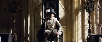 Movie still from “Mary Queen of Scots” (2018), directed by Josie Rourke – A man sitting in a chair wearing a hat; Wide shot, Low angle