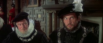 Movie still from “Mary, Queen of Scots” (1971), directed by Charles Jarrott – A man wearing a black hat and a black suit; Close Up shot, Over the shoulder angle