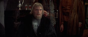 Movie still from “Mary, Queen of Scots” (1971), directed by Charles Jarrott – An old man with a long beard is sitting in front of a wall; Medium shot, Over the shoulder angle