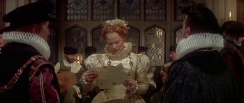 Movie still from “Mary, Queen of Scots” (1971), directed by Charles Jarrott – A woman is holding a piece of paper in front of a group of people; Medium shot, Over the shoulder angle