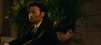 Movie still from “Master Z: The Ip Man Legacy” (2018), directed by Woo-Ping Yuen – A man and a boy sitting next to each other in front of some plants; Close Up shot, Over the shoulder angle