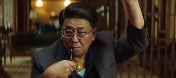 Movie still from “Master Z: The Ip Man Legacy” (2018), directed by Woo-Ping Yuen – An older man wearing glasses and a suit holding a pen in his mouth; Close Up shot, Over the shoulder angle