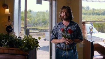 Movie still from “Masterminds” (2015), directed by Jared Hess – A man holding two red roses standing in front of an open door; Medium shot, Over the shoulder angle